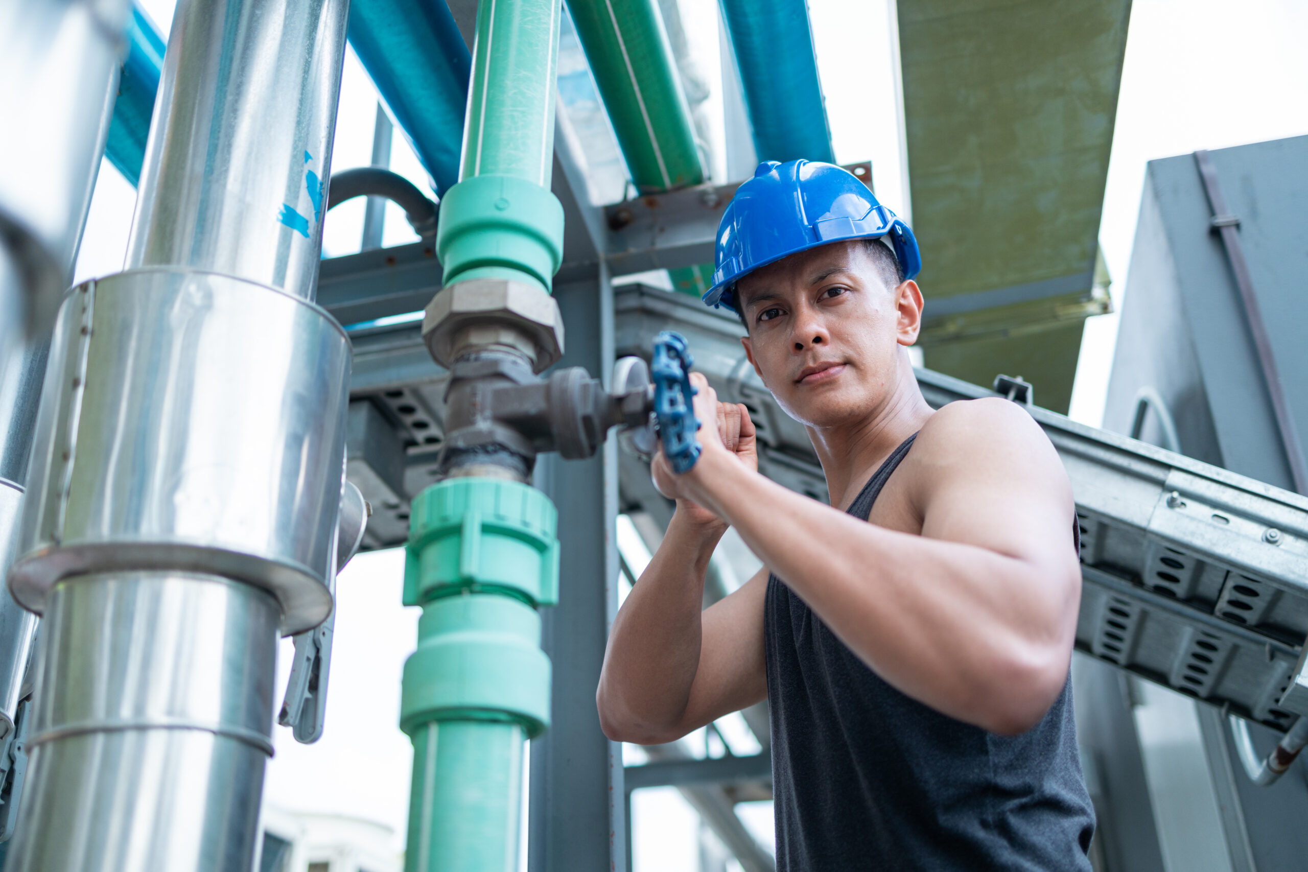 A focused worker in a sleeveless shirt and safety helmet is manually adjusting the valves on an industrial pipeline system.